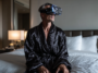 Homme en peignoir de soie sombre assis sur un lit dans une suite d’hôtel de luxe, portant un casque VR, entouré de silhouettes féminines holographiques dans une ambiance nocturne immersive.