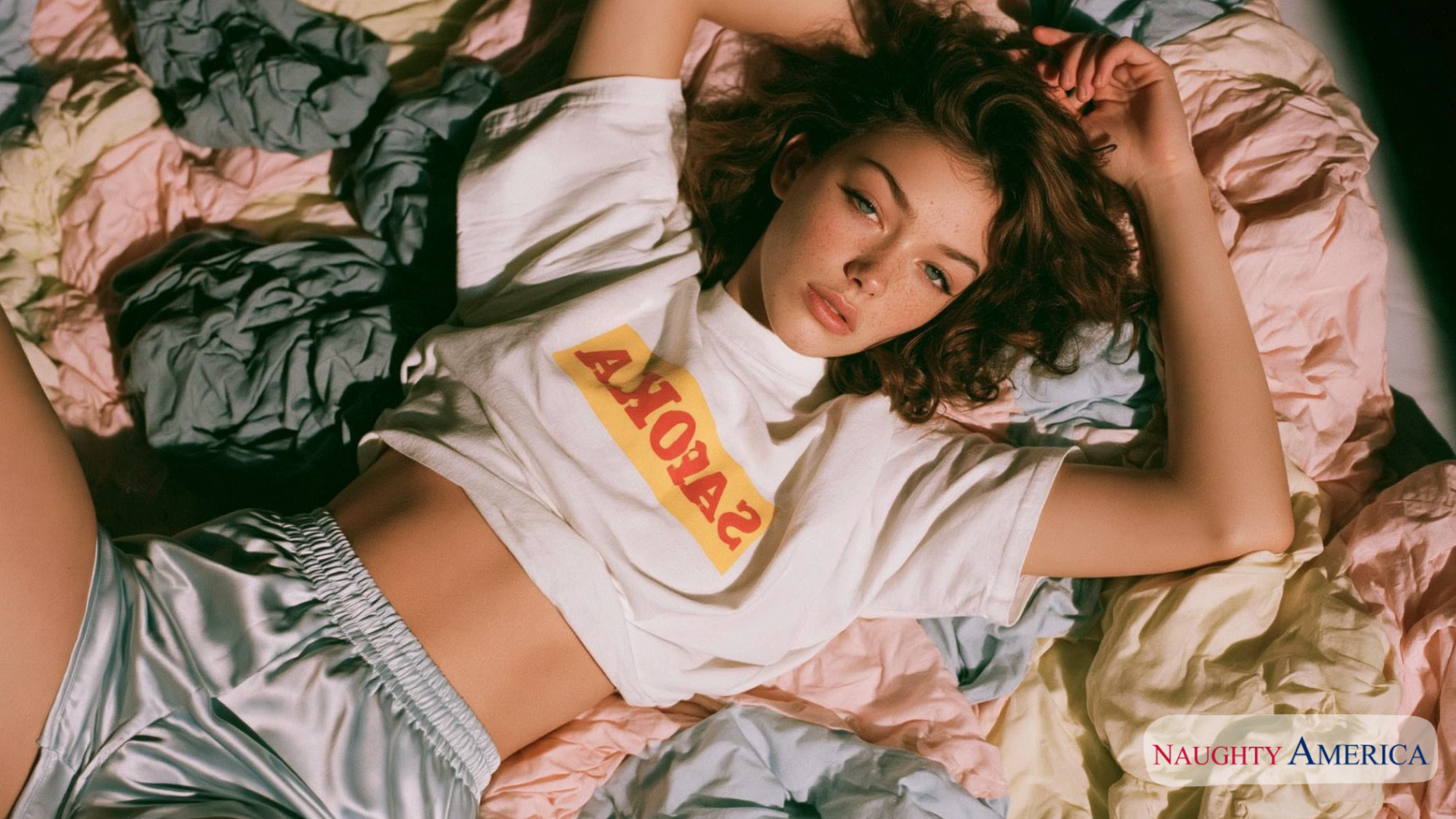 VR porn : Jeune femme aux cheveux bouclés allongée sur un lit, portant un t-shirt blanc court et un short satiné argenté, avec un regard dirigé vers l'objectif.