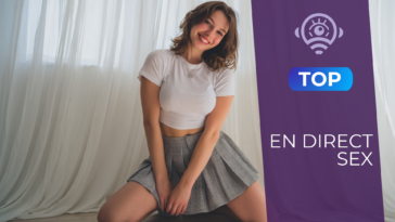 Jeune femme souriante aux cheveux châtains courts, vêtue d’un t-shirt blanc ajusté et d’une jupe plissée grise, prenant la pose face à la caméra lors d’un show de sex en direct.